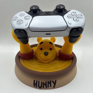 May include: A white and black video game controller resting on a yellow Winnie the Pooh character stand. The stand features a yellow bear head emerging from a yellow honey pot base with the word HUNNY printed in black on the front of the base.