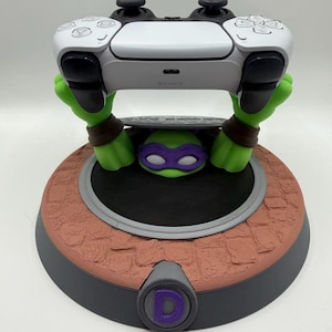May include: A white Sony PlayStation 5 controller resting on a green character stand. The stand features a green figure with a purple mask emerging from a manhole cover base with a textured brown surface and a purple letter D on the front.