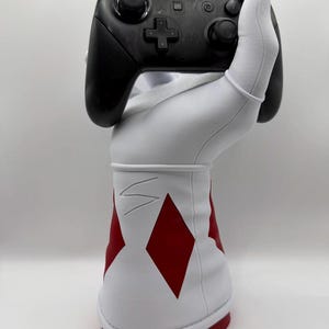 May include: A black Switch Pro controller held by a white and red decorative hand. The hand has a red diamond pattern and a white cuff. The controller is black with various buttons and a directional pad.