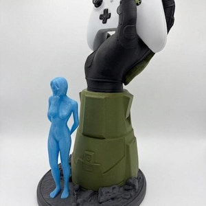 May include: A white Xbox controller is held by a black and green hand-shaped stand. A blue figurine stands to the left of the stand. The base is dark gray with a textured appearance. The stand is olive green.