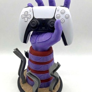 May include: A purple hand-shaped gaming controller stand holding a white and black controller. The stand has a red and purple striped base with gray accents and a circular base. A unique accessory for gamers.