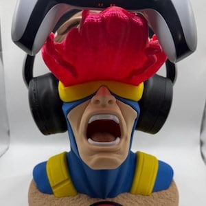 May include: A Cyclops character bust headphone stand featuring a blue and yellow suit, a red translucent visor effect, and a circular base with an X-Men logo. The bust is designed to hold a white and black gaming controller on top and black headphones.