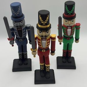 May include: Three colorful nutcracker figurines. The nutcrackers are dressed in blue, red, and green uniforms, each holding a sword. They stand on black bases and have detailed facial features and hats.