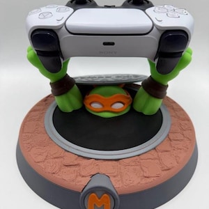 May include: A white and black game controller resting on a green character stand. The stand features a green character with an orange mask emerging from a manhole cover on a textured brown base with a gray circular emblem displaying the letter M.