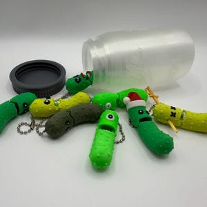 May include: A collection of novelty pickle-shaped keychains in various colors, including green, yellow, and brown, with some featuring cartoon faces and accessories. A clear jar and a black lid are also present.