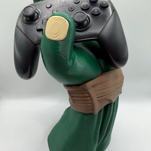 May include: A green sculpture of a hand holding a black Nintendo Switch Pro Controller. The hand has a tan-colored wooden accent on the palm. The controller has black buttons and the word "Switch" printed on it.