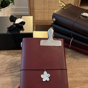 May include: A burgundy leather journal with a silver clip and a flower charm. The journal is held by a person. Other journals and a gift box are in the background.