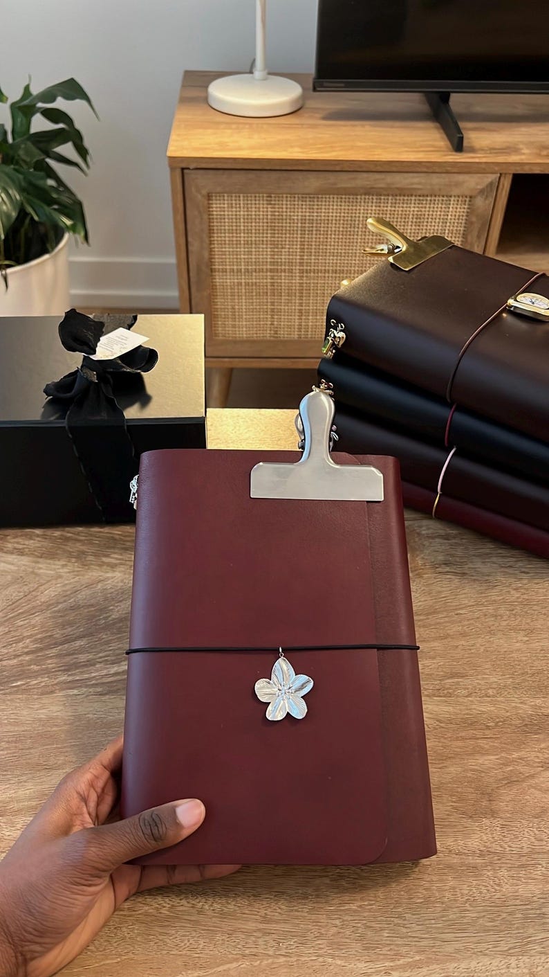 May include: A burgundy leather journal with a silver clip and a flower charm. The journal is held by a person. Other journals and a gift box are in the background.
