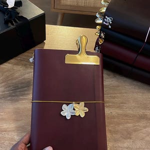 May include: A burgundy leather journal with a gold-colored clip and a decorative floral charm. The journal is secured with a yellow elastic band. Other journals are stacked in the background. The journal is held by a person.