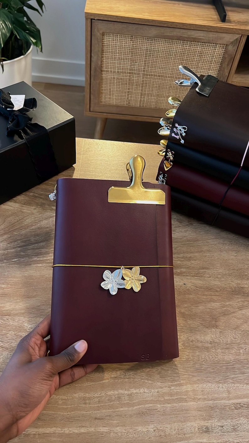 May include: A burgundy leather journal with a gold-colored clip and a decorative floral charm. The journal is secured with a yellow elastic band. Other journals are stacked in the background. The journal is held by a person.