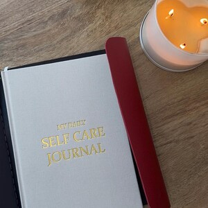 May include: A white journal with gold lettering that reads "MY DAILY SELF CARE JOURNAL" is next to a red bookmark. A lit candle in a heart-shaped glass container is in the background. The journal and bookmark are on a wooden surface.