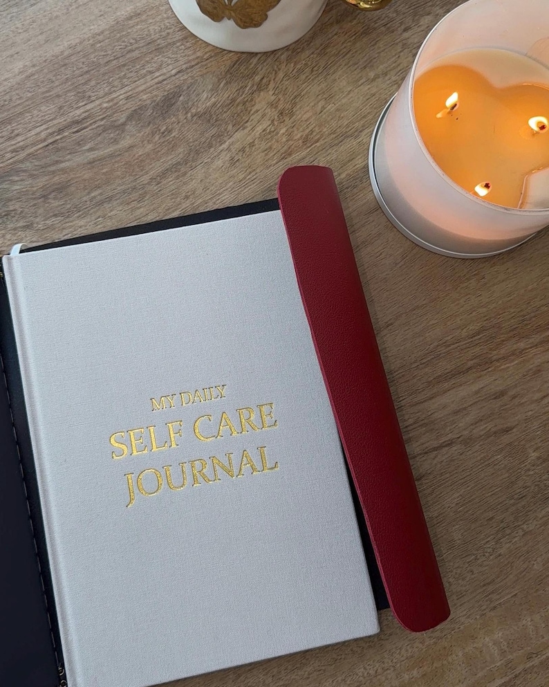 May include: A white journal with gold lettering that reads "MY DAILY SELF CARE JOURNAL" is next to a red bookmark. A lit candle in a heart-shaped glass container is in the background. The journal and bookmark are on a wooden surface.