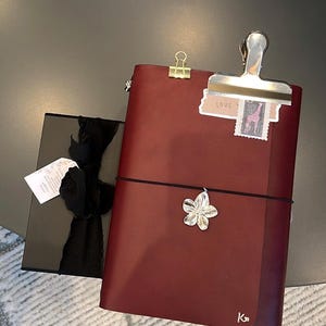 May include: A burgundy leather journal with a black elastic closure and a silver flower charm. A black gift box with a black ribbon is next to the journal. A silver clip holds a small tag that says "LOVE".