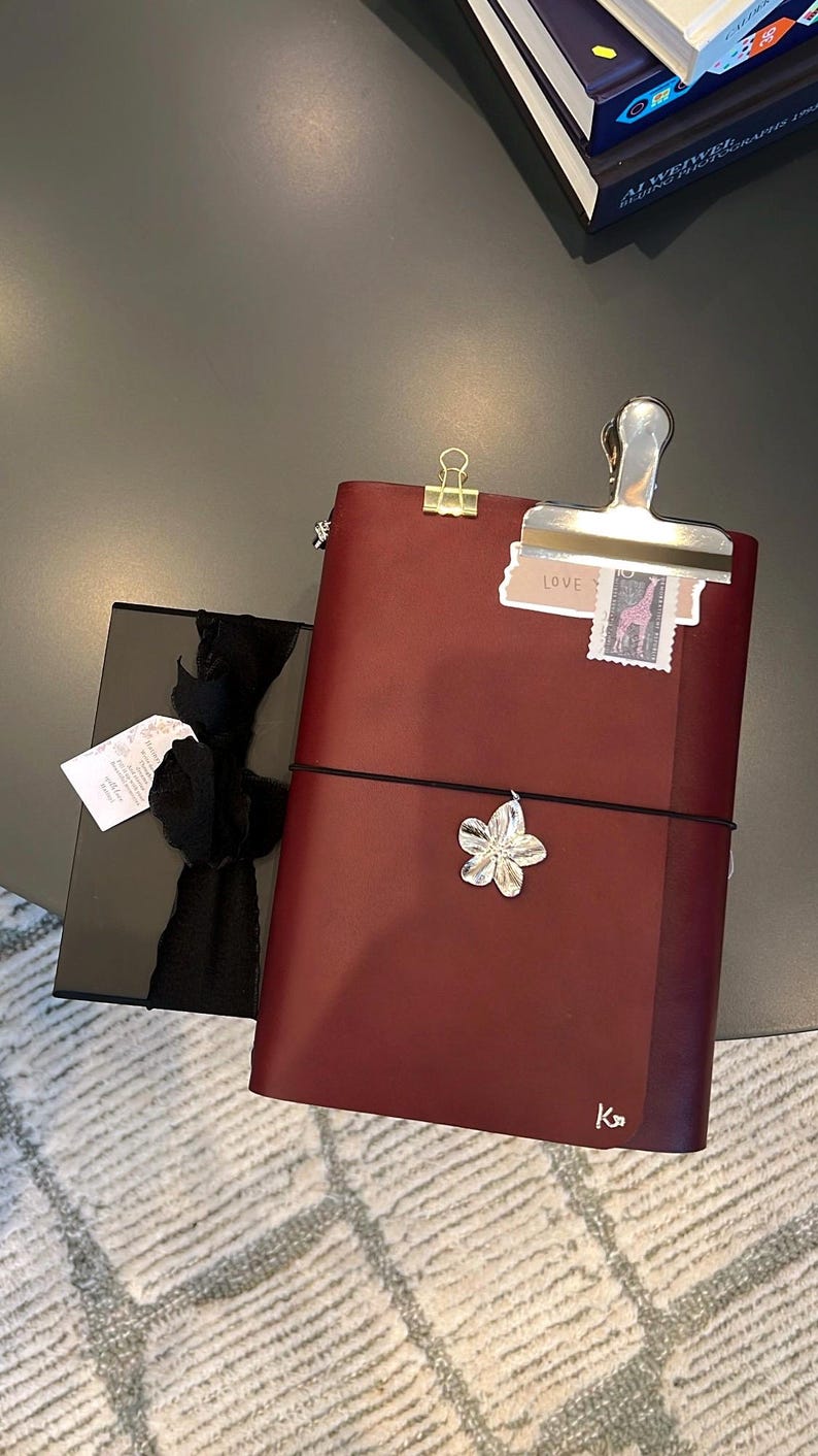 May include: Burgundy leather journal with a black elastic closure and a silver flower charm. A black gift box with a black ribbon is next to the journal. A silver clip holds a small tag that says "LOVE".