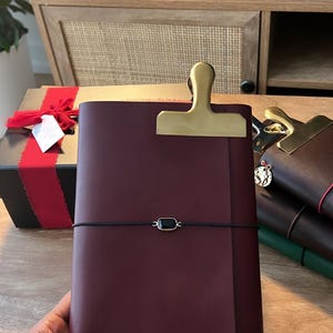 May include: A burgundy leather journal with a gold-colored clip closure and a black elastic band. The journal has the initials "AB" embossed on the lower right corner. Other journals are stacked in the background.