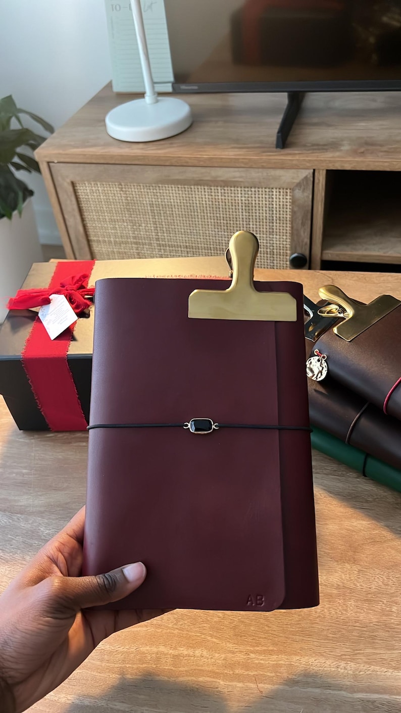May include: A burgundy leather journal with a gold-colored clip closure and a black elastic band. The journal has the initials "AB" embossed on the lower right corner. Other journals are stacked in the background.