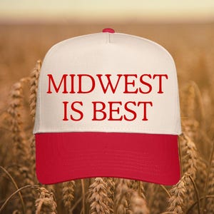 May include: A white and red baseball cap with the text "MIDWEST IS BEST" in red block letters. The cap has a red brim and a red button on top. The background is a field of wheat.