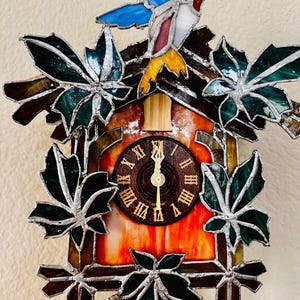 May include: A stained glass cuckoo clock featuring a colorful hummingbird perched atop. The clock face has Roman numerals and gold hands. Green leaves and brown accents frame the clock, creating a decorative and artistic piece.