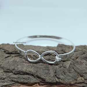 Puede incluir: Un brazalete plateado con un símbolo de infinito en el centro. El brazalete se muestra sobre un trozo de madera marrón oscuro. El nombre de la marca "VinitaSilverShop" es visible en la esquina superior izquierda.