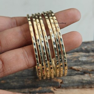 Peut inclure: Ensemble de huit bracelets dorés avec une texture martelée. Les bracelets sont empilés, mettant en valeur leurs motifs individuels. Les bijoux sont présentés sur une surface en bois, soulignant le contraste entre l'or et le bois.