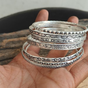 May include: A stack of silver bangles with intricate floral and leaf patterns. The bangles are displayed on a hand, showcasing the detailed designs and the shiny, reflective surface of the metal. The text "VinitaSilverShop" is visible.