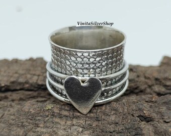 Può includere: Un anello d'argento con una fascia strutturata e un ciondolo a forma di cuore. L'anello ha una texture martellata sulla fascia principale e un cuore liscio e lucido. L'anello è esposto su un pezzo di legno scuro.