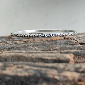 May include: A silver bangle bracelet with an intricate design of repeating oval shapes. The bracelet is displayed on a rough, wooden surface, with the text "VinitaSilverShop" visible on the bracelet.