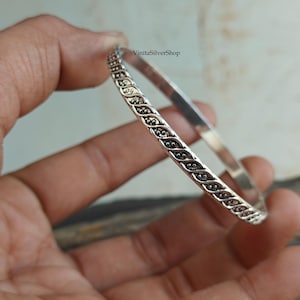 May include: A silver bangle bracelet with an intricate, repeating pattern of curved lines and small, dark dots. The bracelet is held in a hand, and the brand name "VinitaSilverShop" is visible.