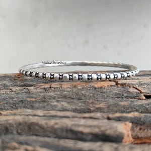 May include: A silver bangle bracelet with a repeating pattern of small, dark square shapes. The bracelet is set against a rustic wooden surface. The text "VinitaSilverShop" is visible on the bracelet.