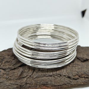 May include: A collection of silver bangles with a textured finish. The bangles are stacked on a piece of dark brown wood. The jewellery is displayed against a white background.