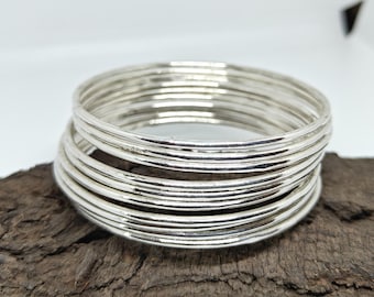 May include: A stack of silver-colored bangles with a textured surface. The bangles are stacked on a piece of dark brown wood. The jewelry is displayed against a white background.
