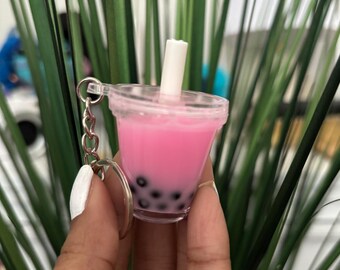 May include: A miniature pink bubble tea keychain with black tapioca pearls and a white straw. The small plastic cup is attached to a silver keyring. The background features green plant leaves.