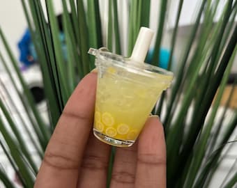 May include: A miniature clear plastic cup filled with yellow liquid and small lemon slices. The cup has a white straw and a clear lid. The cup is held in a hand, with green plant stems in the background.
