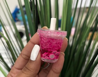 May include: A miniature pink bubble tea cup with a white straw. The cup is filled with pink liquid and small, clear, cube-shaped pieces. Dragon fruit slices are visible at the bottom of the cup. The cup is held in a hand.