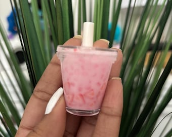 May include: A miniature clear plastic cup filled with a pink beverage and small red pieces, resembling a drink. A white straw is inserted into the drink. The cup is held in a hand with a white painted fingernail, with green plants in the background.