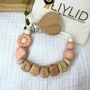 Könnte beinhalten: Ein personalisierter Schnullerhalter für Babys mit dem Namen "LUISA" aus Holzbuchstaben. Der Clip hat ein Holzherz, eine rosa Blume und rosa und beige Silikonperlen. Im Hintergrund befindet sich ein kleiner Beutel mit dem Text "LIYLID".