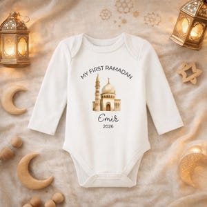 Könnte beinhalten: Weißer Baby-Langarmbody mit dem Text "MY FIRST RAMADAN" über einer Aquarell-Illustration einer Moschee. Der Name "Emir" und das Jahr "2026" sind darunter gedruckt. Der Body ist von dekorativen Elementen umgeben.