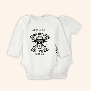 Peut inclure: Deux bodies blancs pour bébés. L'un a des manches longues, l'autre courtes. Les deux ont un graphisme noir d'un crâne avec un chapeau de paille et les mots "NEW TO THE STRAW HAT CREW" et "ONE PIECE" en texte noir.