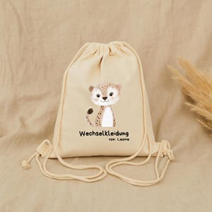 Pode incluir: Uma mochila bege com cordão com uma ilustração de chita de desenho animado. A chita tem manchas marrons e está sorrindo. O texto "Wechselkleidung" e "von: Leonie" são impressos abaixo.