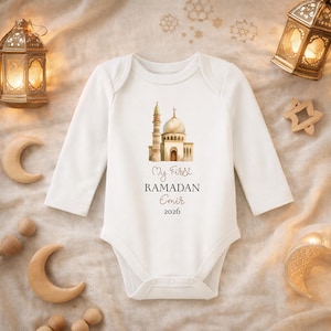 May include: A white baby onesie with long sleeves featuring a watercolor illustration of a mosque. The text "My First Ramadan Emir 2026" is printed below the image. The onesie is laid flat on a beige surface.