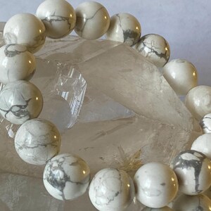 May include: A white howlite bead bracelet. The round beads have gray veining patterns. The bracelet is displayed on a clear, crystalline surface.