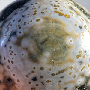 May include: Close-up of a polished orb featuring a unique pattern. The surface displays a mix of white, gray, and brown hues, with small, circular, golden-brown spots scattered across the surface. The orb has a smooth, glossy finish.