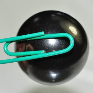 Puede incluir: Una esfera negra con un clip de papel verde sujeto a ella.