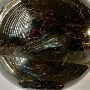 Puede incluir: Una esfera oscura y pulida con una superficie reflectante. La esfera tiene inclusiones internas plumosas y un sutil brillo iridiscente. El color general es un negro profundo con toques de marrón y azul.