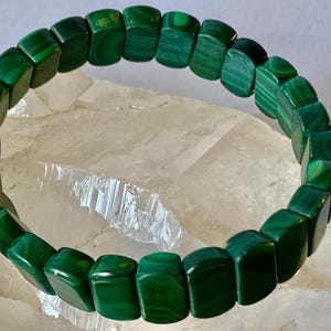 May include: A vibrant green malachite bracelet. The bracelet is composed of rectangular, polished beads with natural patterns. The beads are strung together to form a circular shape. The bracelet is displayed on a light-colored, crystalline surface.