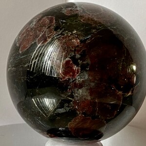 May include: A polished black sphere with red and green inclusions. The sphere is made of a natural stone and has a smooth, shiny surface.