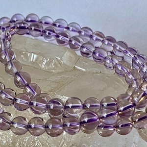 May include: Strands of amethyst gemstone beads. The beads are round, translucent, and a soft purple color. The beads are strung together, creating a necklace or bracelet. The background is a light, textured surface.