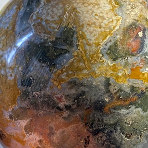 May include: A polished, spherical gemstone with a complex pattern of colors. The surface displays a mix of dark gray, olive green, tan, and reddish-brown hues, with areas of white and gold. The stone's texture appears smooth and reflective.