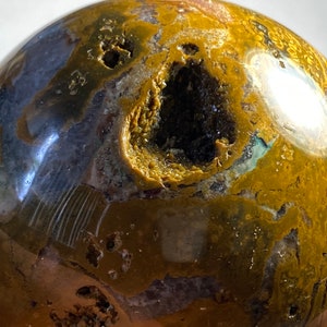 May include: A polished, spherical gemstone with a complex pattern of brown, yellow, and blue hues. The orb features a cavity with a dark interior, adding depth and visual interest. The surface has a glossy finish.