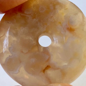 May include: A round, light brown agate pendant with a hole in the center. The agate has a swirling, cloudy pattern in shades of white and brown.